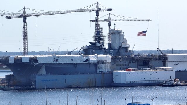 Navy ship docked at Norfolk Naval Shipyard at Portsmouth, Virginia undergoing repairs and updates during peacetime with American flag waving in breeze 