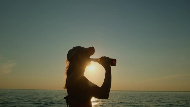 Silhouette of a young long haired woman drinking from a bottle by the sea at sunset. A refreshing sip of beer on a hot summer evening on vacation.