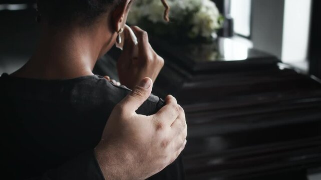 Close-up over the shoulder shot of tearful African American woman comforted by unrecognizable male hand, standing in front of coffin at funeral