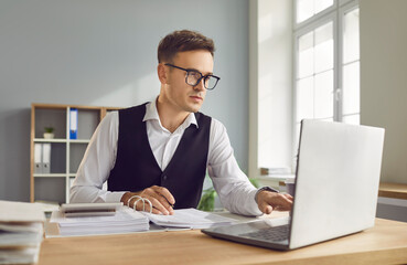 Obraz premium Portrait of young attractive business man accountant working at the desk with a pile of financial documents on his workplace looking at the laptop monitor screen at the office and typing.