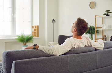 From behind man in casual wear, with smart wrist watch sitting on gray sofa in modern white living...