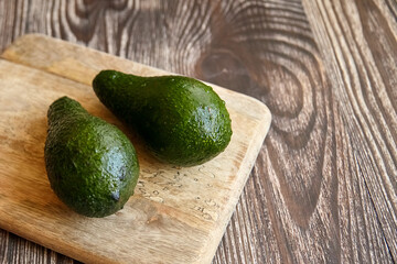 Close-up of two wet green avocados placed on a rustic wooden cutting board, set against a dark wood grain surface with natural light and texture detail