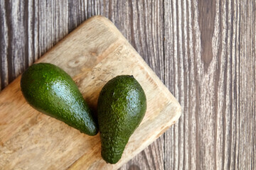 Two ripe green avocados placed on the corner of a light wooden cutting board against a rustic dark wood grain surface, photographed in natural lighting, top view