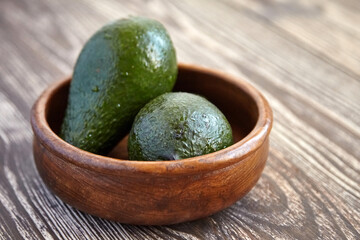Two ripe green avocados placed inside a smooth round wooden bowl resting on a rustic wooden table with visible texture and soft natural lighting