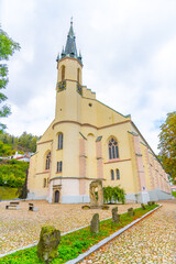 Naklejka premium The Church of St. Joachim stands elegantly in Jachymov, Czechia, showcasing its architectural beauty amidst lush greenery and cloudy skies. Visitors admire the historic structure.