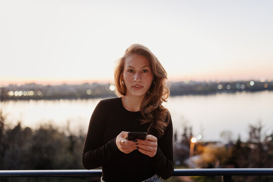 Woman holding smart phone while standing on balcony overlooking river at sunset