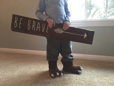 Close-up of a boy stand-in in a house wearing bear slippers and holding a "Be Brave" sign