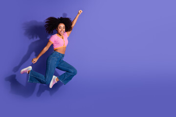 Smiling young woman in casual attire joyfully jumping against a vibrant purple background,...