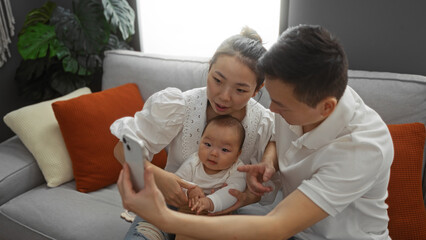Chinese family with mother, father, and baby taking a selfie indoors in a cozy living room setting on a sofa, capturing a moment of love and togetherness.