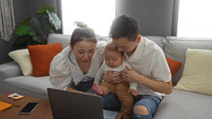 Family with mother, father, and baby boy sitting on sofa in living room looking at laptop together, showcasing love, warmth, and togetherness in a cozy home environment.