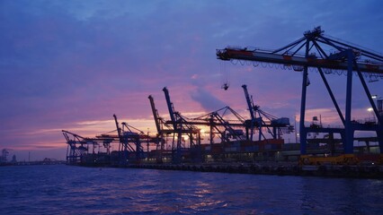 Obraz premium A breathtaking view of idle shipping cranes at a port during sunset, with calm waters reflecting vibrant sky colors and creating a tranquil ambiance.