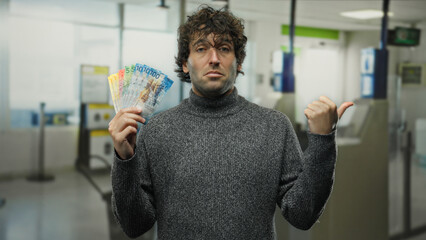 Hispanic man indoors at airport terminal holding swiss franc banknotes while pointing aside,...
