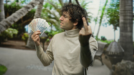 Hispanic man outdoors in park holding polish zloty banknotes with an angry expression and middle finger gesture, surrounded by blurred natural greenery.
