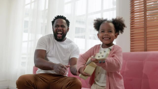 enjoy happy love black family african american father playing ukelele with his daughter, smiling and having fun moments good time on pink sofa in the white living room at home. father day concept.