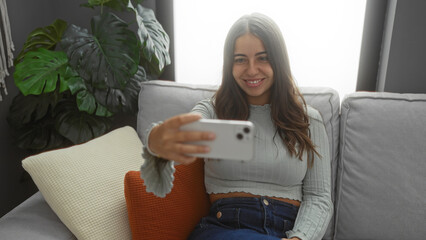 Obraz premium Woman smiling while taking a selfie in a cozy living room with lush green plants, showcasing hispanic beauty and comfort in an indoor home setting.