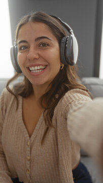 Woman smiling in living room wearing headphones, taking selfie indoors, young brunette female enjoying music in cozy apartment environment.