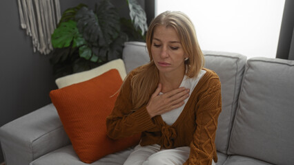 Fototapeta premium Woman experiencing discomfort indoors sitting in a cozy home living room wearing a brown cardigan with orange pillow and green plant in the background.