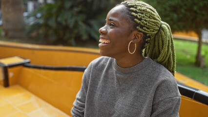 Woman with braids smiling outdoors in an urban park setting, showcasing casual fashion and joy in a...