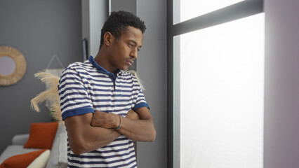 Young man with crossed arms looks through a window with a sad expression in a cozy living room,...