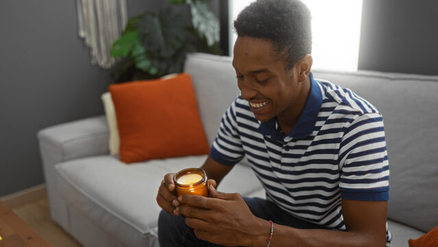 Young man relaxing indoors smiling and holding a lit candle, sitting on a comfortable sofa in a stylish living room apartment with warm decor and natural lighting.