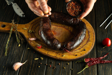 A pair of hands adds seasoning to a dark sausage resting on a wooden board. Surrounding ingredients...