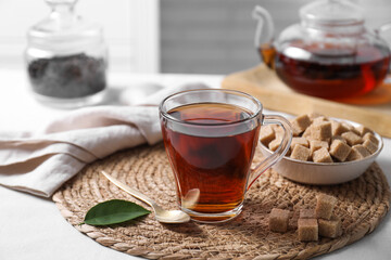 Aromatic black tea in cup and brown sugar cubes on white table, closeup