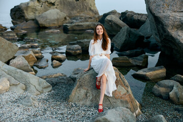 Woman in white dress with red shoes sitting on ocean rock, contemplating the vast sea