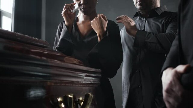Man comforting crying African American woman, dabbing her eyes with tissue, as they standing in dark clothing beside wooden coffin at funeral