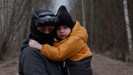 A young man in a mask with FPV glasses on his head, holding his son in his arms,