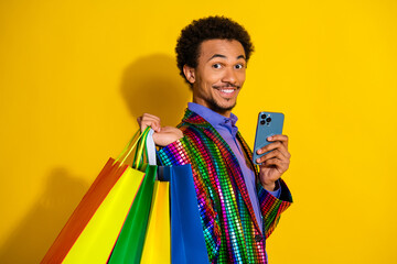 Charismatic young man holding colorful shopping bags and a smartphone against a vibrant yellow...