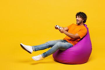 Full body young Indian man wear orange t-shirt casual clothes sit in bag chair hold in hand play pc...