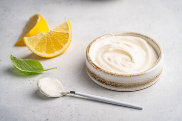 Bowl of tasty sour cream or Greek yogurt with a spoon and lemon slices on white background, closeup