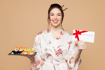 Young satisfied woman she wear kimono clothes hold eat raw fresh sushi roll served on black plate gift certificate coupon voucher card isolated on plain beige background studio. Japanese food concept.