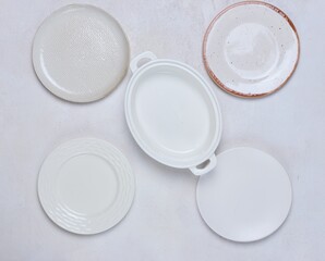 Four white empty ceramic plates of different shapes and baking dish on light concrete background. Tableware, serving items.