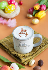 A cup of hot chocolate with mousse of milk ,latte art in bunny shape draw on it and festive Easter decoration with chocolate Easter eggs and tulip flowers.