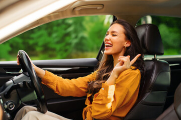 Excited woman driving her new car and enjoying listening to music, holding auto steering wheel and...