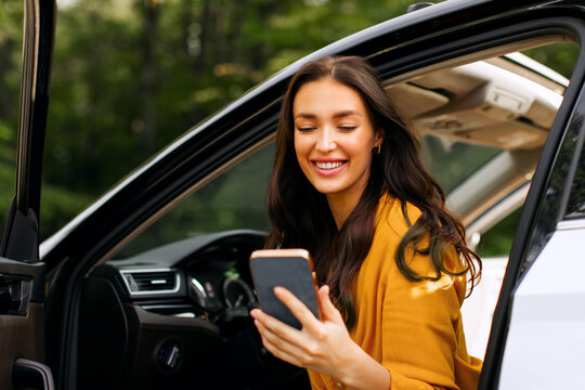 Positive woman getting out of her car and using cellphone, sitting with opened door, stopping for break of road or checking navigator app