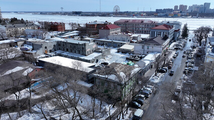 Winter aerial view of a city with a mix of old and modern buildings, snowy streets, and parked cars. Some buildings appear under renovation. Bare trees and snow-covered rooftops highlight the cold.
