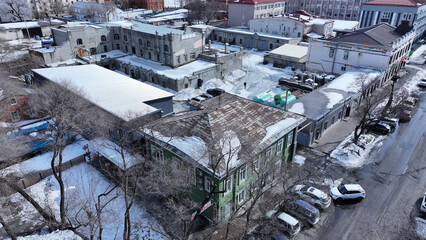 Winter aerial view of a city with a mix of old and modern buildings, snowy streets, and parked cars. Some buildings appear under renovation. Bare trees and snow-covered rooftops highlight the cold.