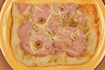A piece of pizza with mortadella and pine nuts on a yellow plate, shot from above on a wooden...
