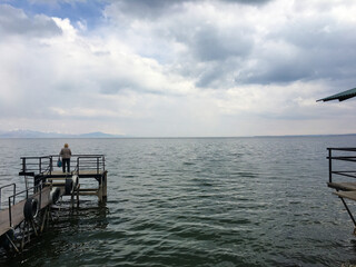 Wooden pier extending into lake beneath cloudy sky and surrounded by forest Single figure stands at dock’s edge looking over tranquil water and hazy horizon