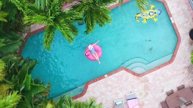 Top view of vibrant pool surrounded by palm trees, with elegant woman relaxing on pink flamingo float. Aerial swimming pool in tropical garden Miami, Florida. Woman in pool during summer vacation 4K