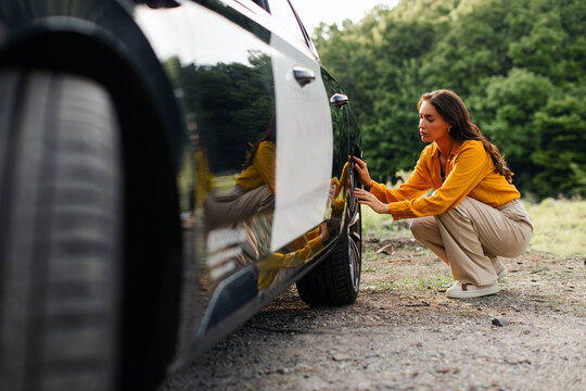 Young woman broken down on country road, sitting near rear wheel and checking car tire for puncture, having problem