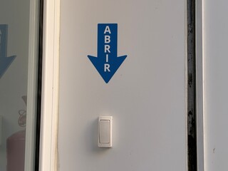 A blue sign with the word abrir and a downward arrow is mounted on a white wall beside a doorbell. This simple design clearly communicates the action to open the door