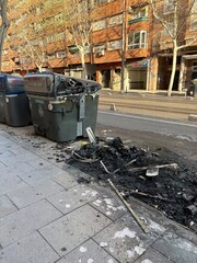 A city street shows a damaged trash bin, charred and smoldering, with burnt debris scattered on the sidewalk, indicating recent fire activity. Nearby buildings provide urban context