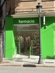 A vibrant green pharmacy features a clear entrance displaying various health products. Sunlight illuminates the cheerful ambience along the pedestrian street, encouraging visitors to stop by