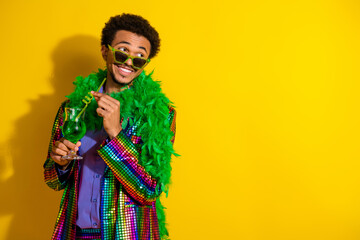 Young stylish man in vibrant outfit and boa holding a cocktail against a bright yellow background showcasing party vibes.