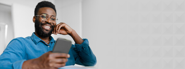 A young African American manager is smiling while using his smartphone at the office. He is engaged and relaxed, showcasing success and a modern lifestyle in a professional environment, copy space