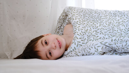Rolled up person. A child wrapped in a blanket with sticking out head lies on the bed in the bedroom. Sunny morning dream. Psychology comfort. Happy smiling caucasian boy. High quality photo.