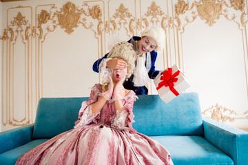 Elegant couple in baroque costumes enjoying a surprise gift exchange on an ornate sofa in a vintage...